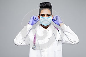 Portrait of a handsome indian doctor in scrubs with stethoscope and mask standing confidently , isolated on white background