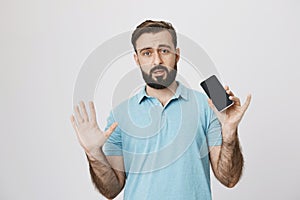Portrait of handsome male with a beard holding smartphone and his palms up while standing next to the white wall. Man