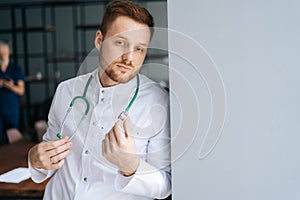 Portrait of handsome male doctor wearing white medical uniform standing with stethoscope in hospital office near window