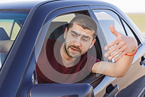 Portrait of handsome male driver waves from car window, being stuck in traffic jam, feels nervous and worried, looks into distance