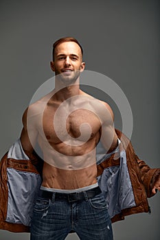 Portrait of a handsome male model posing shirtless in a brown leather jacket and jeans. Grey background. Fashionable