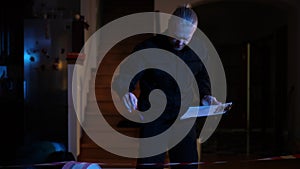 Portrait of handsome male police officer filling in report standing in darkness in robbed house. Confident concentrated