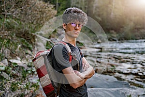 Portrait of handsome male traveler with backpack at fast river in mountains. Wild nature walk.