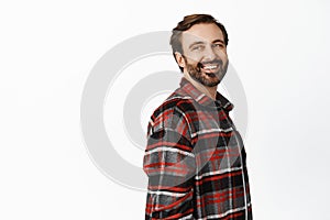 Portrait of handsome man with beard, 35 years old, looking behind at promo empty space, smiling and laughing, standing