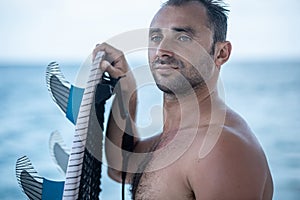 Portrait of handsome man with blue eyes, surfer holding surf board with blue fins and blue ocean