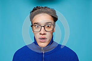 portrait of a handsome man in a blue zip-up sweater and black eyeglasses, standing on a light blue background looking at