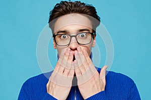 portrait of a handsome man in a blue zip-up sweater and black eyeglasses, standing on a light blue background looking