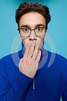 portrait of a handsome man in a blue zip-up sweater and black eyeglasses, standing on a light blue background looking