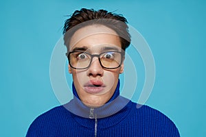 portrait of a handsome man in a blue zip-up sweater and black eyeglasses, standing on a light blue background looking at
