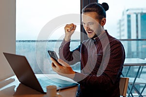 Portrait of handsome man feeling happy, doing winner gesture, video chatting on smartphone with the client, working at laptop,