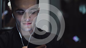 Portrait of handsome man in headphones listening to music and browsing on mobile phone in public transport. City lights