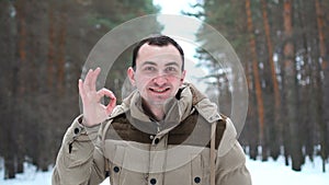 Portrait of handsome man in jacket is showing okay gesture at camera. Man stands in winter forest