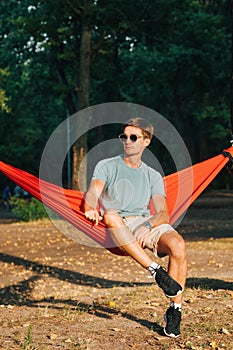 Portrait of a handsome man in light casual clothes resting sitting on a hammock in the park at sunset, wearing glasses, looking