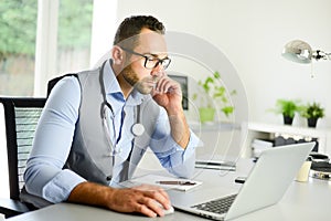 Portrait of handsome man male doctor in medical practice office writing prescription in laptop computer