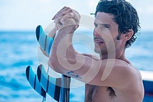 Portrait of handsome man, shirtless surfer with tattoo anchor and surf board with blue fins, blue ocean on background in Maldives