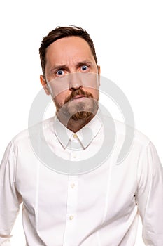 Portrait of handsome man with stubble has indignant expression, frowns face, dressed in white shirt, isolated over white