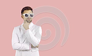 Portrait handsome man in stylish sunglasses wearing in classic white suit and pink shirt, keeps hand touching his chin