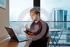 Portrait of handsome man working at laptop, holding smartphone and cup of espresso coffee in hands, background of panoramic