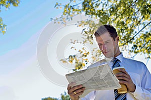 Portrait of handsome mature businessman holding cup of coffee while reading newspaper in park during break