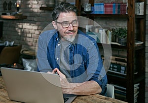 Portrait of handsome mature man with grey hair, wearing red glasses, working on laptop, sitting at desktop at home or in the