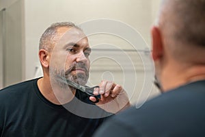 Portrait of handsome mature man trimming his beard himself standing in bathroom