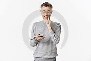 Portrait of handsome middle-aged man in glasses and gray sweater, looking tempted at delicious cake, wanting to eat