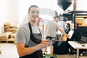 Portrait of handsome modern man wearing apron use phone while standing at coffee roasting machine in local roastery