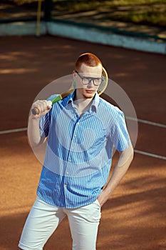 Portrait of handsome redhead young man in stylish casual clothes and glasses posing on opendoor tennis court with tennis
