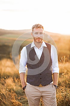 Portrait of handsome retro stylish man, posing on the background of summer field, sunset. Young bearded blond man