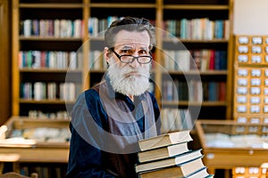 Portrait of handsome senior bearded retired man, librarian or teacher, choosing books in library, holding stack of books