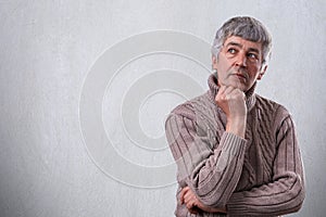 A portrait of handsome senior man standing near white wall dreaming about something looking aside holding his hand under his chin.