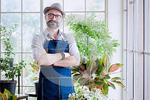 Portrait of a handsome senior man standing and wearing a denim apron is happily taking care of the plants in the nursery.