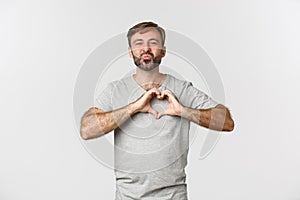 Portrait of handsome smiling man with beard, showing heart sign, confess in love, kissing someone, standing over white