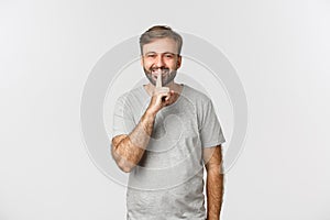 Portrait of handsome smiling man in grey t-shirt, hushing at you kindly asking to keep quiet, standing over white