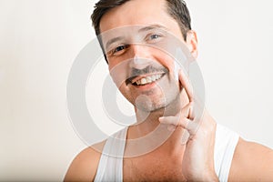 Portrait of handsome smiling toothy man applying moisturizer to his cheek, softening face cream and looking at camera indoors.