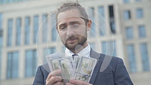 Portrait of handsome successful businessman counting pack of money outdoors. Happy confident Caucasian man looking at
