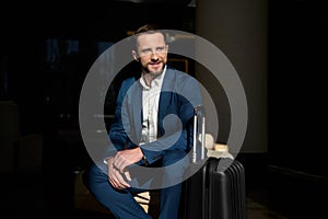 Portrait of a handsome successful man, confident businessman, smiling investor sitting in a chair in a hotel lobby with a luggage