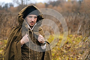 Portrait of handsome traveler man wearing green raincoat tent standing in thicket of bushes in cold overcast day and