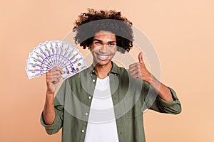 Portrait of handsome trendy cheerful wavy-haired guy holding 100 us banknotes showing thumbup isolated on beige pastel