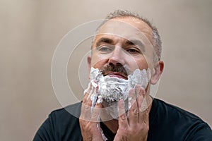 Portrait of handsome white-haired beared middle-aged man applying shaving foam to trim his beard