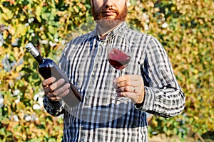 Portrait of handsome wine maker holding in his hand bottle and a glass of red wine and tasting it, checking wine quality while