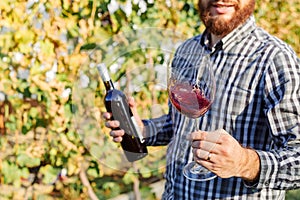 Portrait of handsome wine maker holding in his hand bottle and a glass of red wine and tasting it, checking wine quality while