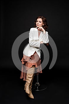 Portrait of handsome woman in long dress and boots sits on a chait and poses for the camera, picture isolated on black