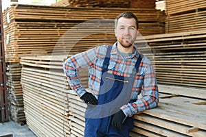 Portrait of a handsome worker choosing the best wooden boards. Carpenter standing next to a big stack of wood bars in a