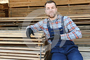 Portrait of a handsome worker choosing the best wooden boards. Carpenter standing next to a big stack of wood bars in a