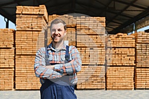 Portrait of a handsome worker choosing the best wooden boards. Carpenter standing next to a big stack of wood bars in a