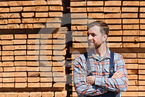 Portrait of a handsome worker choosing the best wooden boards. Carpenter standing next to a big stack of wood bars in a