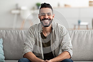 Portrait of handsome young Arab man smiling and looking at camera, sitting on sofa at home