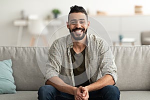 Portrait of handsome young Arab man smiling and looking at camera, sitting on sofa at home