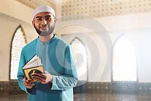 Portrait of handsome young asian muslim man with beard posing, hold holy book quran